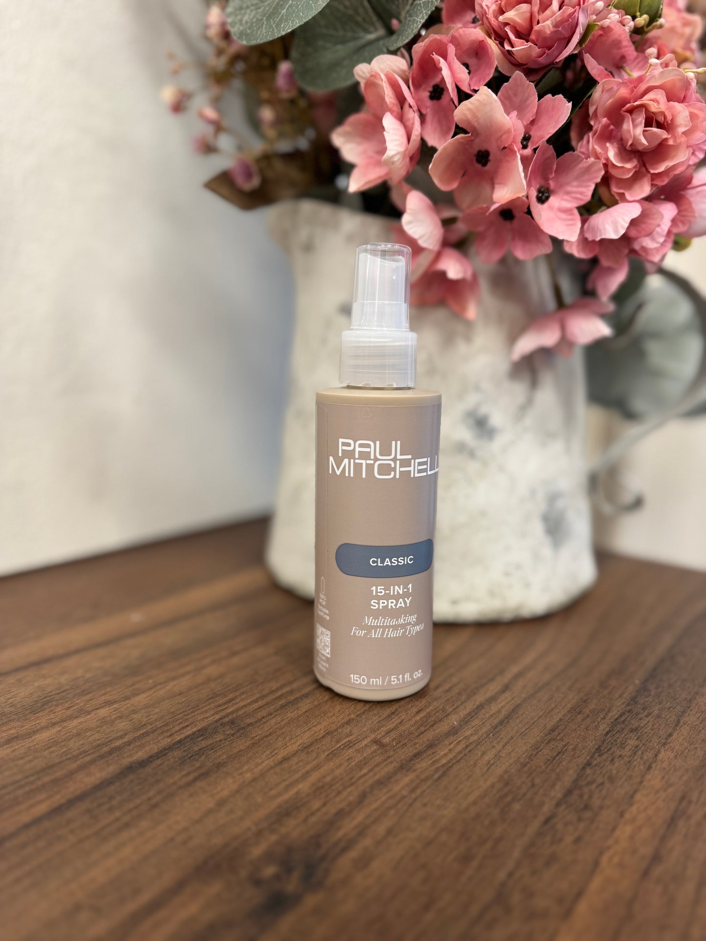 Paul Mitchell 15-in-1 Spray Leave-in Pflege, braune Flasche auf Holztisch vor Blumen – Paul Mitchell