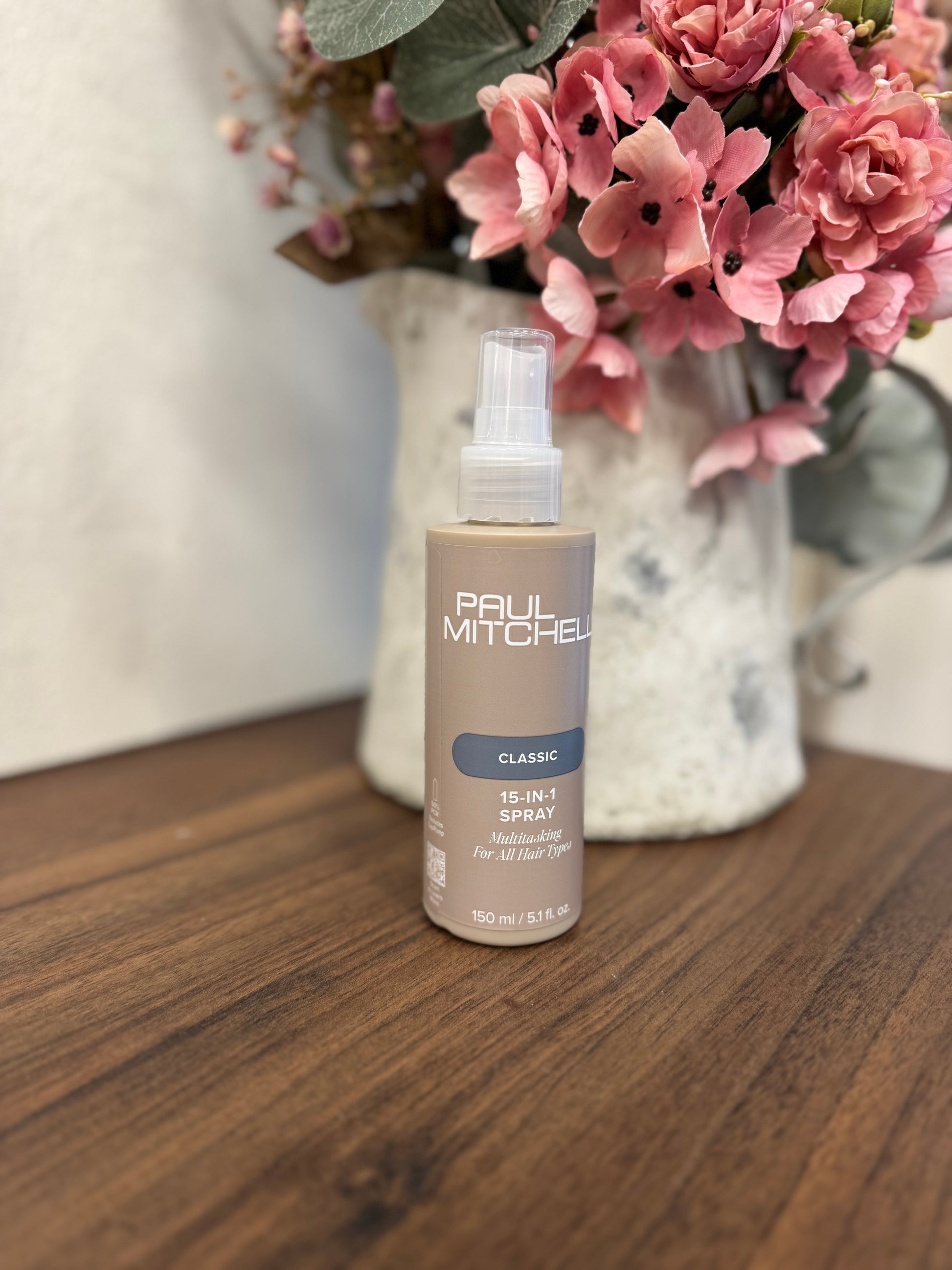 Paul Mitchell 15-in-1 Spray Leave-in Pflege, braune Flasche auf Holztisch vor Blumen – Paul Mitchell