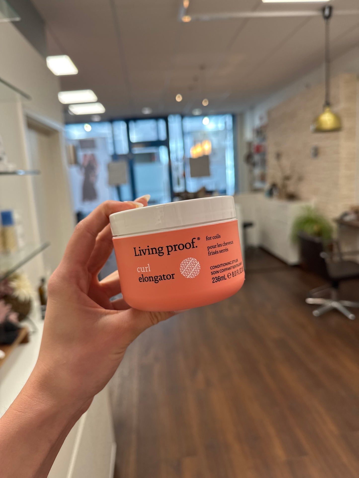 Living Proof Curl Elongator Lockencreme, Dose in Hand im Friseursalon mit unscharfem Hintergrund – Living Proof