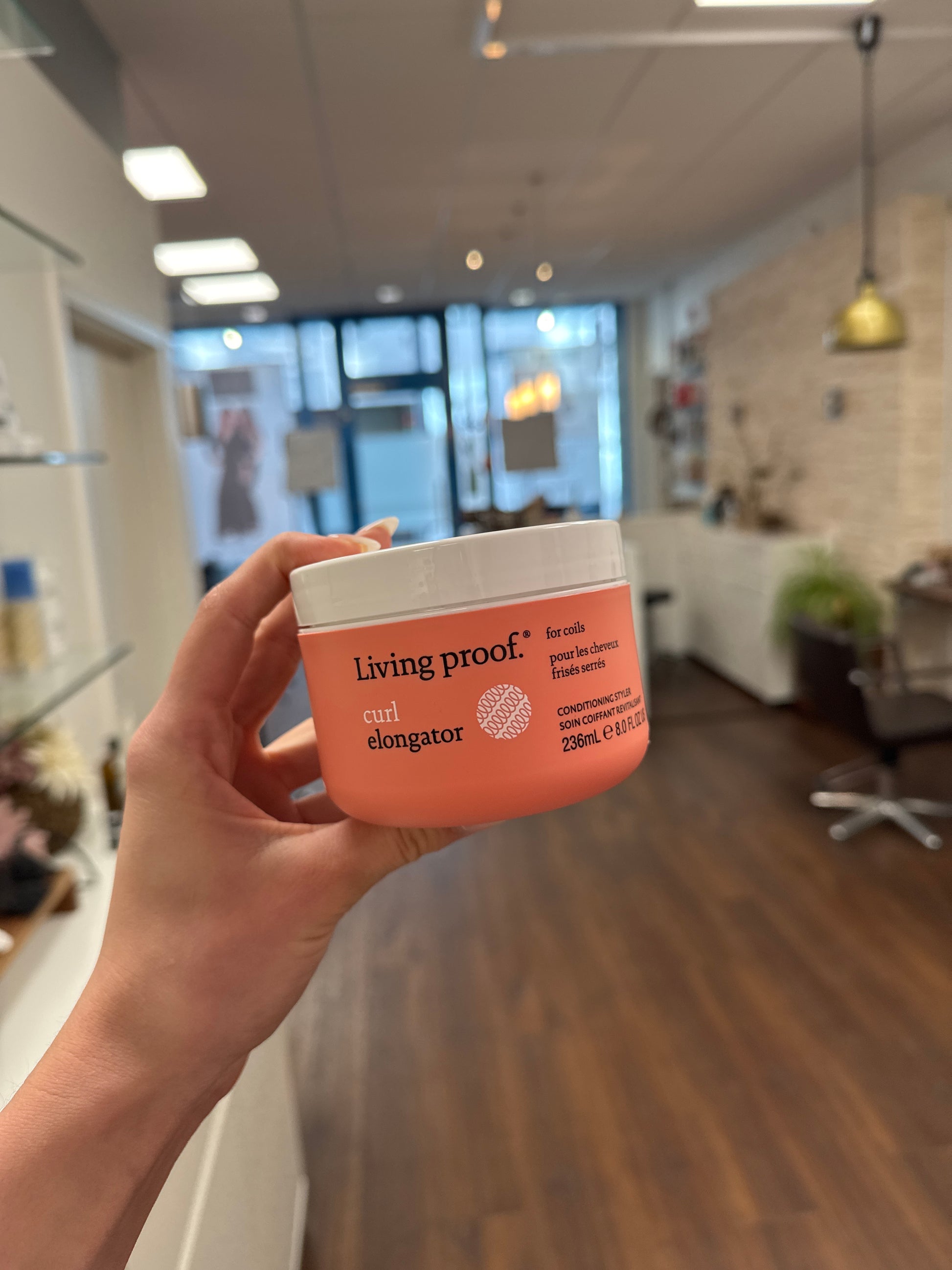Living Proof Curl Elongator Lockencreme, Dose in Hand im Friseursalon mit unscharfem Hintergrund – Living Proof