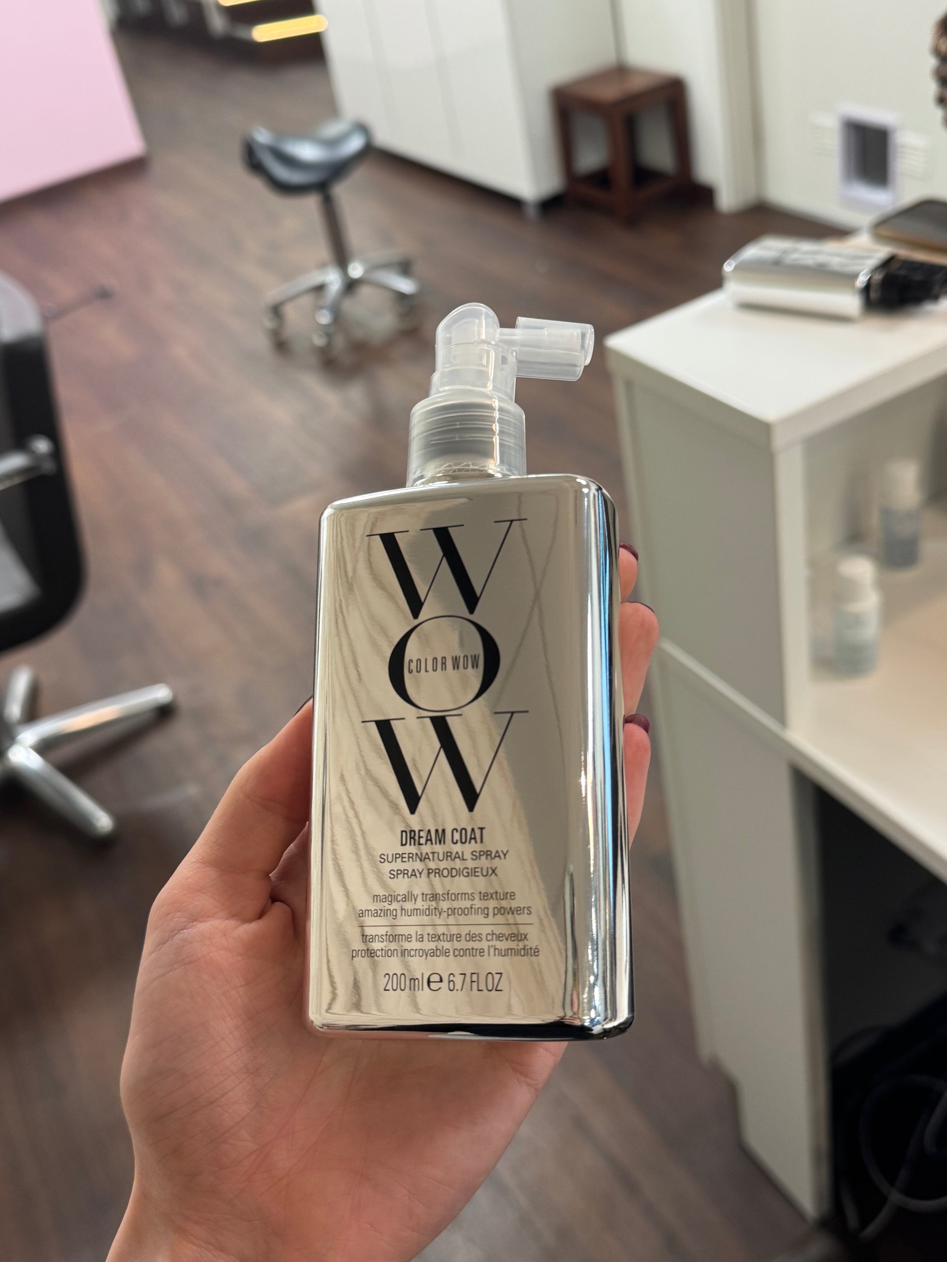 Color Wow Dream Coat Supernatural Spray in der Hand gehalten im Friseursalon mit Arbeitsbereich im Hintergrund – Color Wow