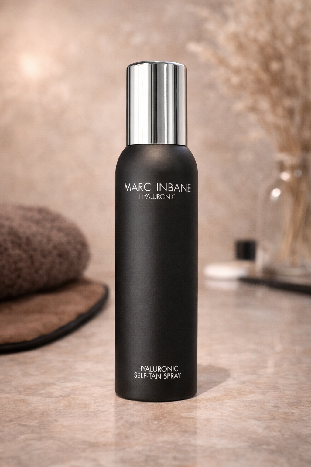 Marc Inbane Hyaluronic Self Tan Spray für gleichmäßige Bräune, Produkt stehend auf heller Steinfläche mit Handtuch und Pflegeaccessoires – Marc Inbane
